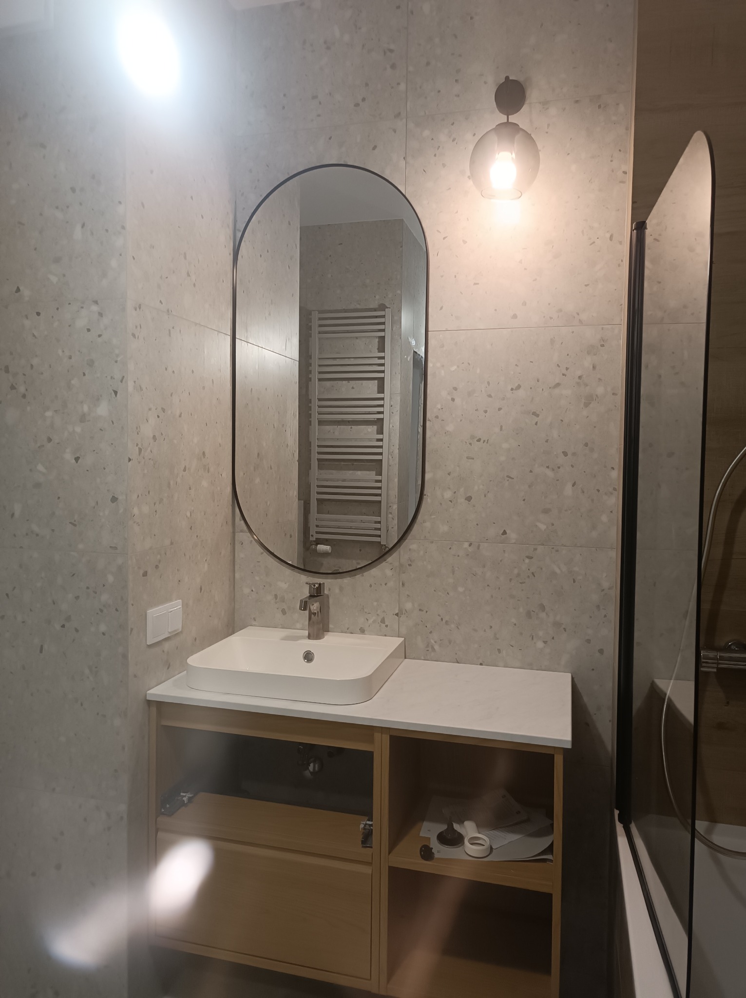 Jasna łazienka z owalnym lustrem, umywalką na szafce z drewnianymi frontami i szklanym panelem prysznicowym. Ściany wyłożone płytkami imitującymi lastryko.