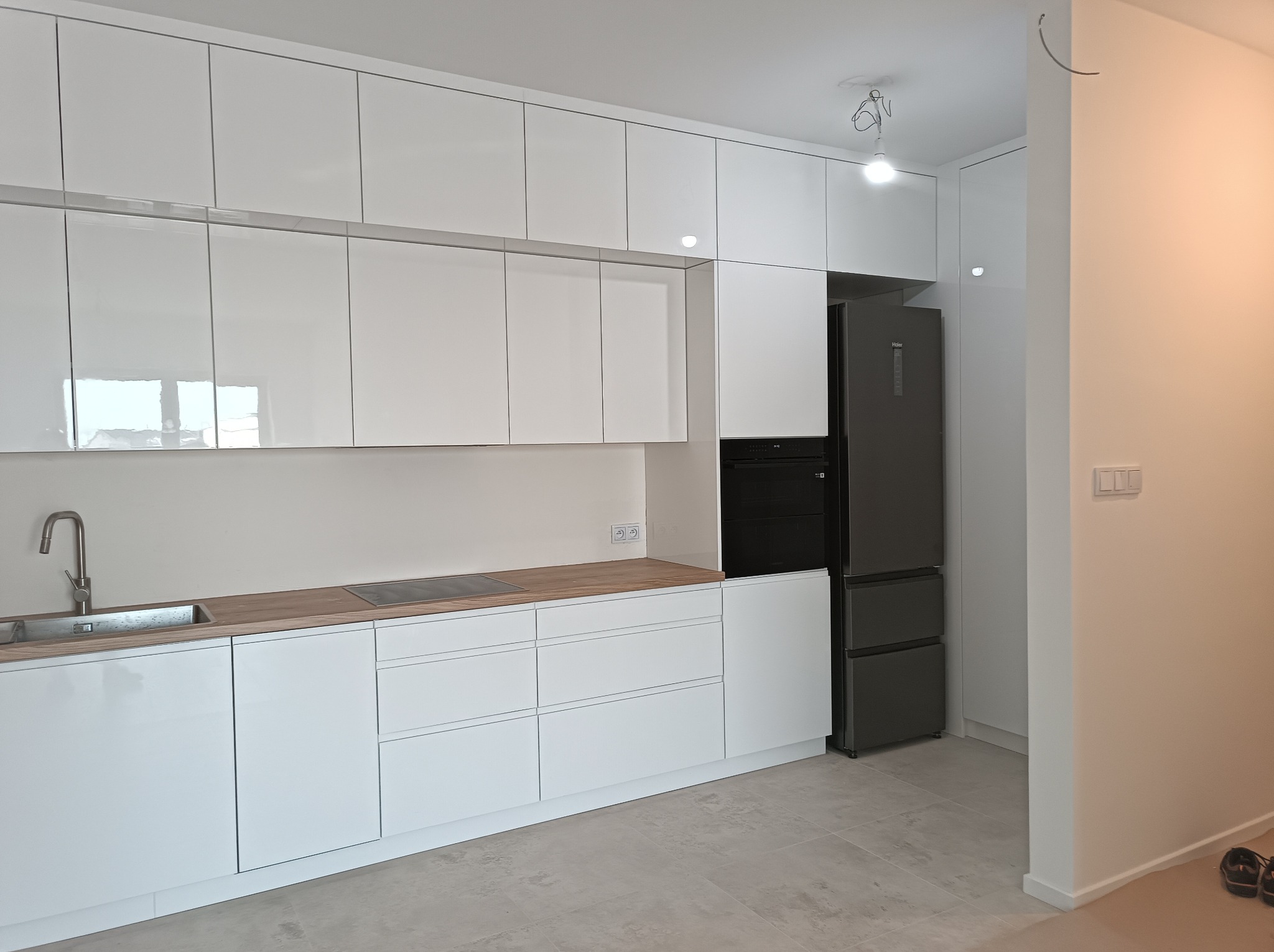 Biała, nowoczesna zabudowa kuchenna z drewnianym blatem, czarnym piekarnikiem i lodówką. Minimalistyczny design, jasne wnętrze, szara podłoga.