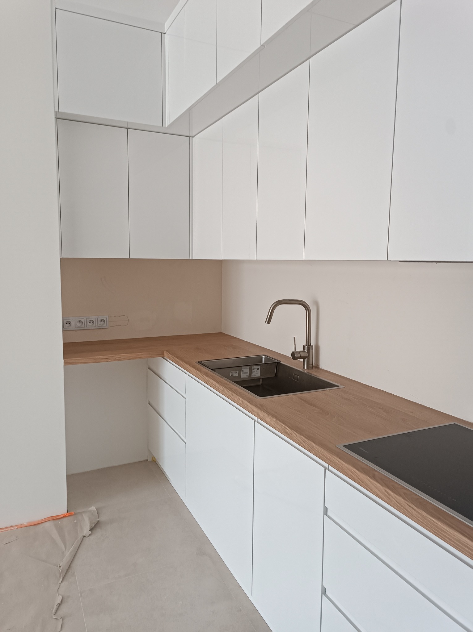 Nowoczesna kuchnia z białymi szafkami i drewnianym blatem. Widoczny zlew, płyta indukcyjna i gniazdka elektryczne. Minimalistyczny design, jasne wnętrze.