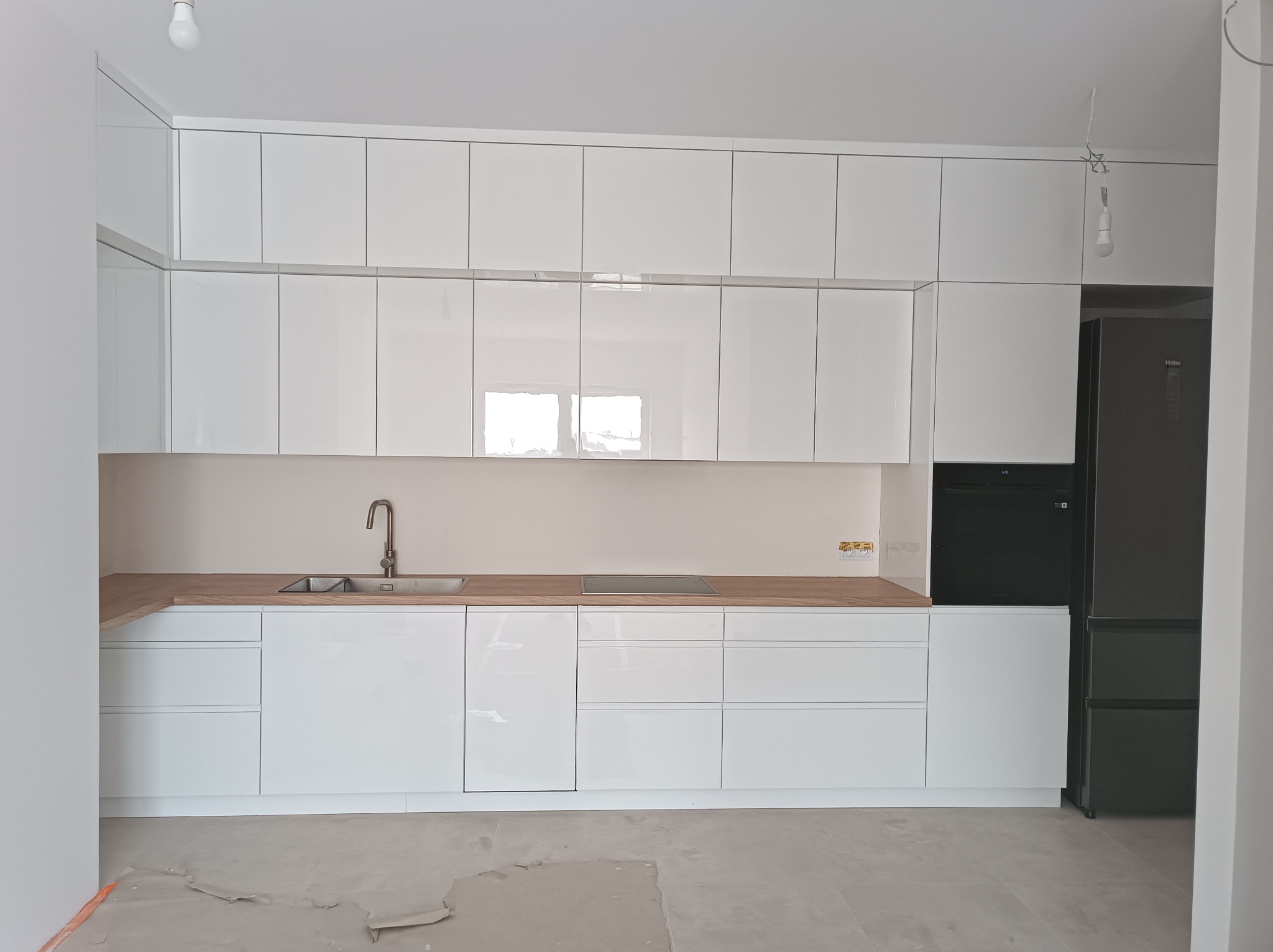 Biała, nowoczesna zabudowa kuchenna z drewnianym blatem i czarnym sprzętem AGD. Widoczne niedokończone wykończenie podłogi i wisząca żarówka.