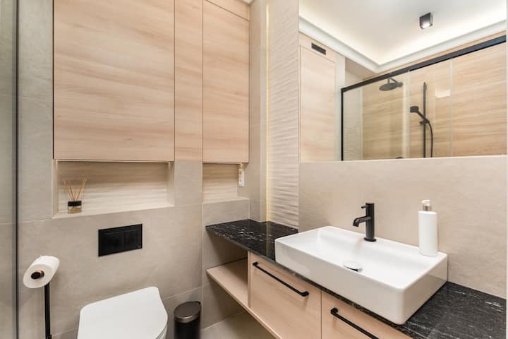 Nowoczesna łazienka z beżowymi płytkami, drewnopodobnymi szafkami, czarnym blatem z umywalką nablatową i kabiną prysznicową w tle.