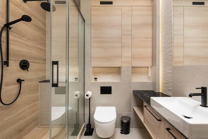 Nowoczesna łazienka z kabiną prysznicową typu walk-in, toaletą, umywalką nablatową i szafkami w jasnym drewnie, armatura w kolorze czarnym.