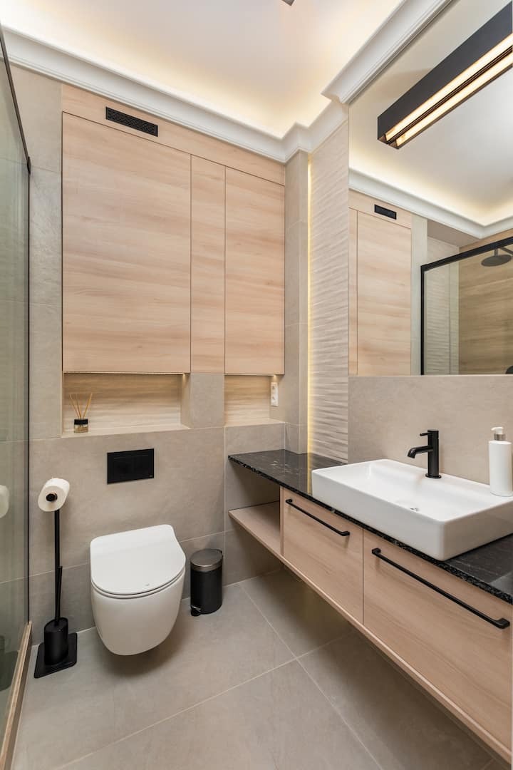 Nowoczesna łazienka z beżowymi płytkami, drewnianymi szafkami, czarnym blatem i armaturą, podświetleniem LED oraz wiszącą toaletą z czarnym przyciskiem.