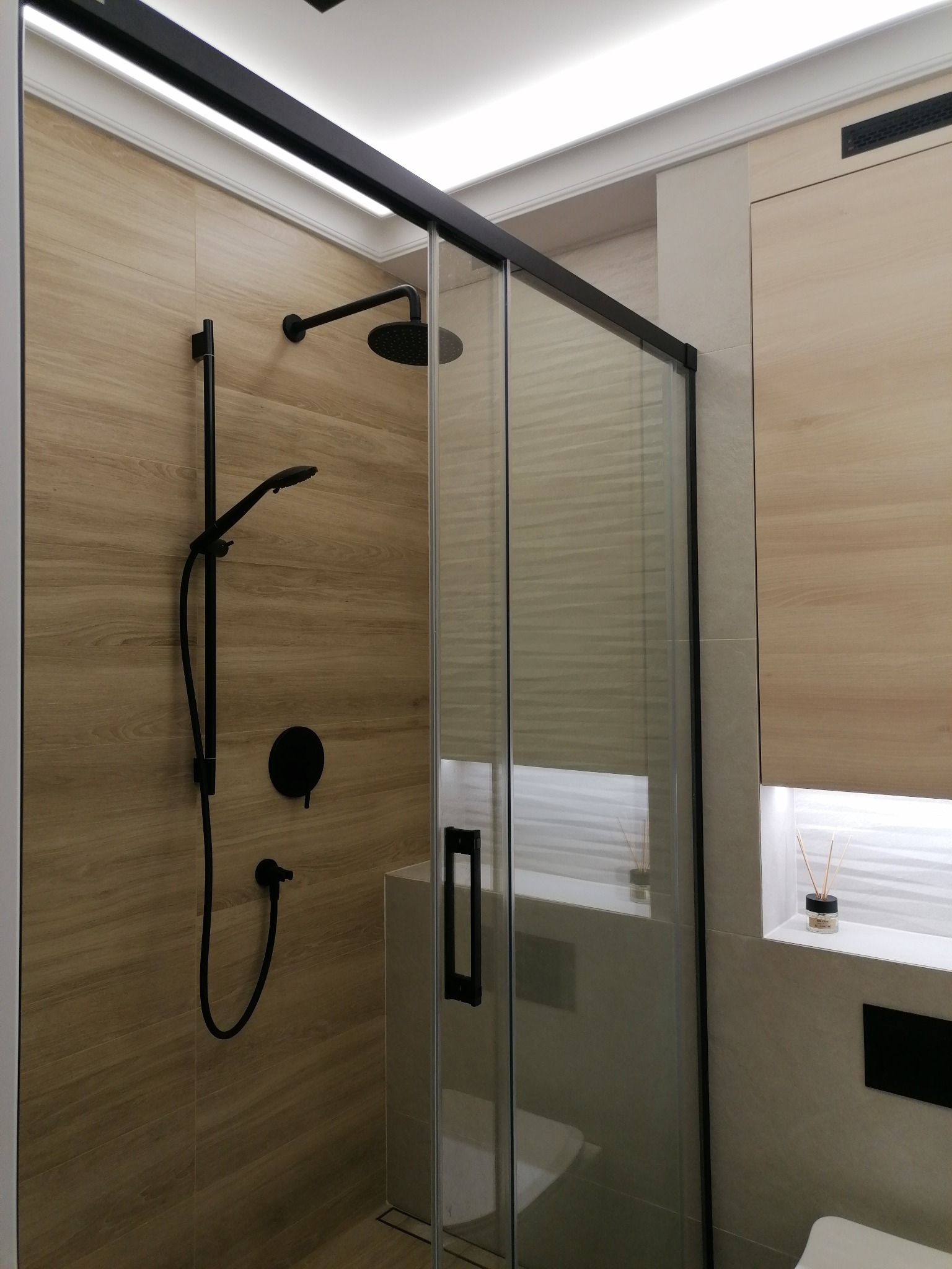 Nowoczesna kabina prysznicowa z czarnymi elementami armatury na tle płytek imitujących drewno, minimalistyczny design z oświetleniem LED podsufitowym.