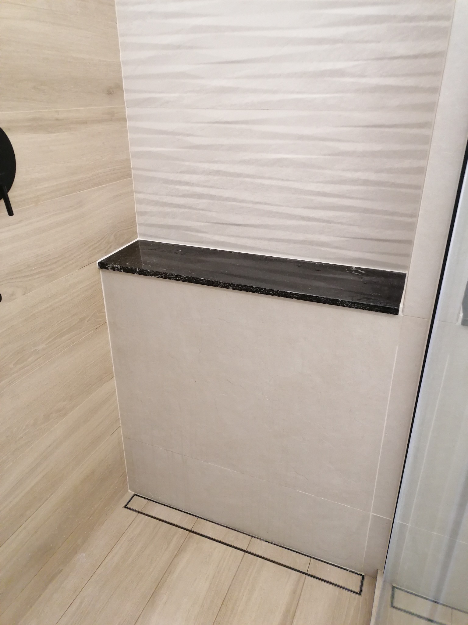 Wykończony fragment łazienki z beżowymi płytkami imitującymi drewno, czarnym odpływem liniowym i półką z czarnego kamienia w strefie prysznicowej, płytki ścienne z delikatnym wzorem fal.