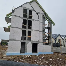 Wznoszony budynek z betonu komórkowego w stanie surowym otwartym, z widoczną konstrukcją schodów zewnętrznych, na tle błotnistego terenu i częściowo wykończonego domu sąsiedniego.