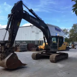 Ciężka, czarno-żółta maszyna budowlana Volvo z łyżką, zaparkowana na placu przed budynkiem z logo Volvo, widoczna w słoneczny dzień.