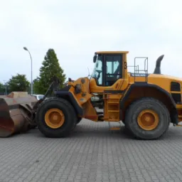 Żółta ładowarka kołowa Volvo L180G zaparkowana na szarym brukowanym placu, widok z boku, z widocznym dużym łyżką i numerem '3' na kabinie.