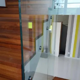 Szklana balustrada zamocowana na drewnianych schodach w nowoczesnym wnętrzu z paskowaną ścianą. Widoczne metalowe mocowania szkła.