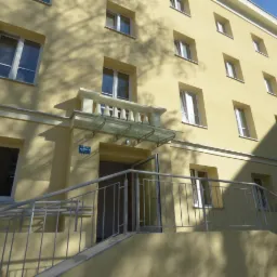 Wejście do budynku z żółtą elewacją, zadaszone szklaną konstrukcją wspartą na kolumnach, z metalową balustradą prowadzącą po schodach.