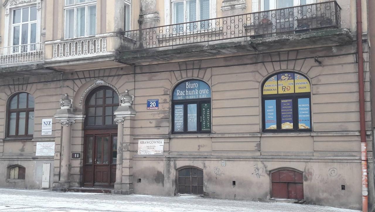 Fasada kamienicy z biurami: rachunkowym i doradztwa finansowego. Widoczne okna z reklamami usług finansowych i rachunkowych, tabliczka z adresem i numerem budynku oraz szyld krawcowni.