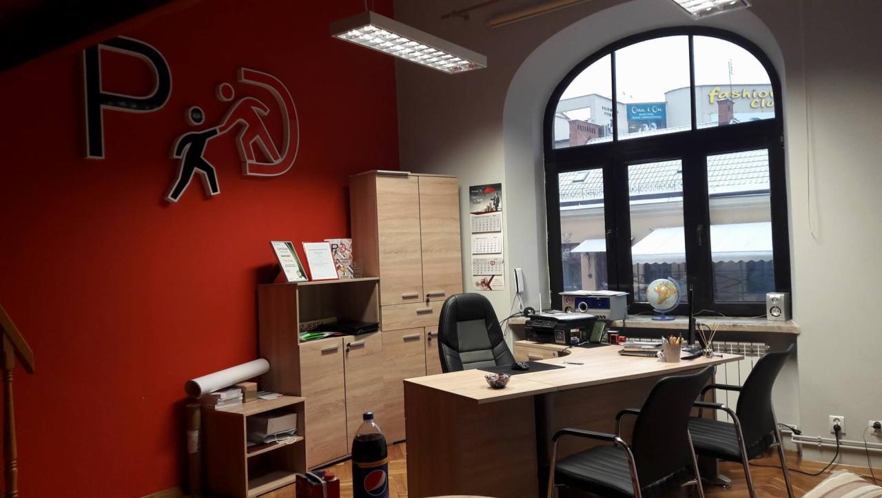 Wnętrze biura z czerwonym akcentem na ścianie, logo firmy, biurko z krzesłami, szafa z dokumentami i widok na ulicę przez duże okno.