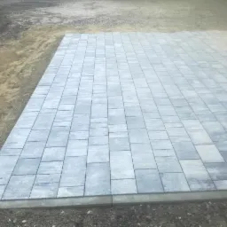 Świeżo ułożona, szara kostka brukowa o prostokątnym kształcie, widoczna na tle przygotowanego podłoża z piasku i żwiru.