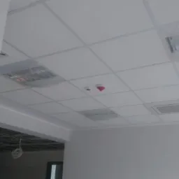 Biały sufit podwieszany kasetonowy z kratką, zabezpieczone folią lampy i czujnik dymu na tle jasnych ścian w pomieszczeniu biurowym.