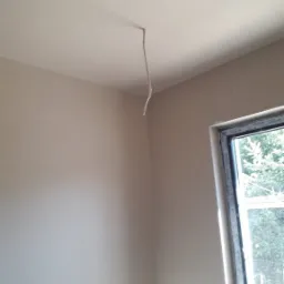 Świeżo położony, jasny tynk na ścianach i suficie w rogu pokoju z wystającym kablem elektrycznym z sufitu oraz oknem z widokiem na zieleń.