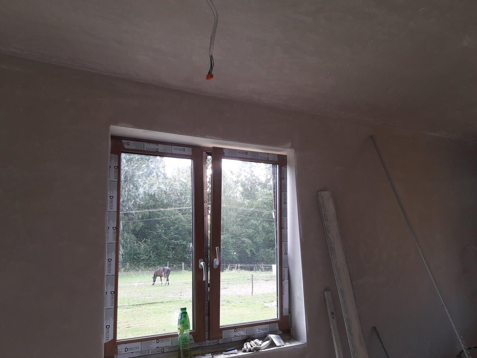 Świeżo otynkowane ściany w pokoju z oknem na pastwisko, gdzie pasie się koń; widoczne wystające kable i butelka na parapecie.