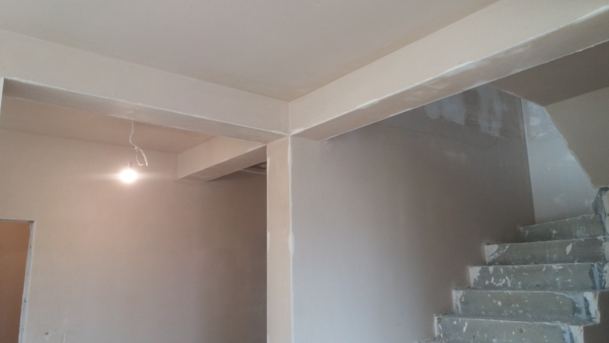 Świeżo położone tynki maszynowe na ścianach i suficie w surowym stanie, widoczne niedokończone schody i wisząca żarówka, ukazujące etap prac wykończeniowych.