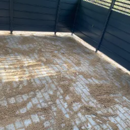 Świeżo ułożona kostka brukowa z piaskiem do wypełnienia szczelin, otoczona nowoczesnym, czarnym ogrodzeniem panelowym w słoneczny dzień.