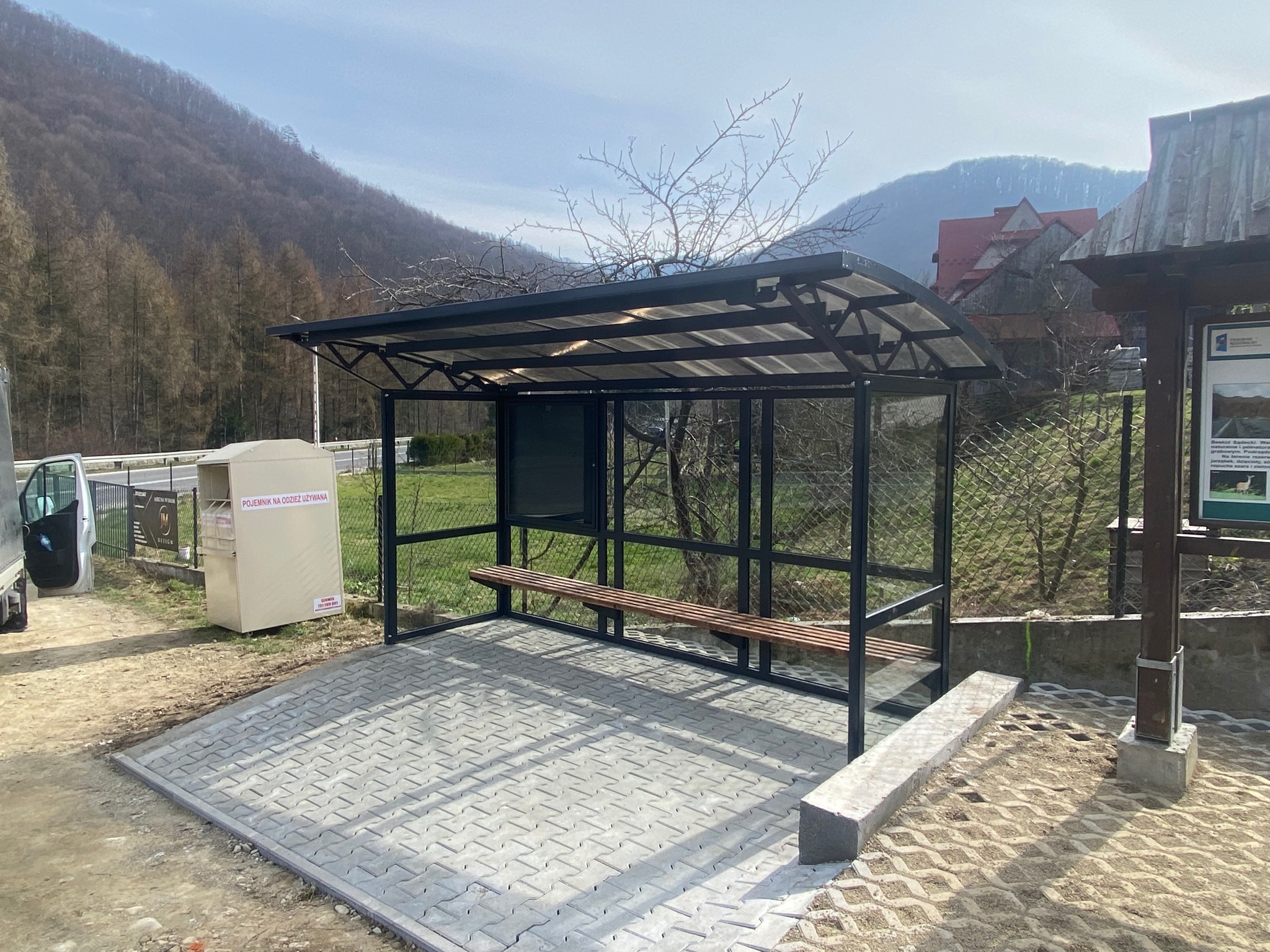 Nowoczesna wiata przystankowa z drewnianą ławką i szarą kostką brukową, z widokiem na góry w tle, obok kontener na odzież używaną i tablica informacyjna.