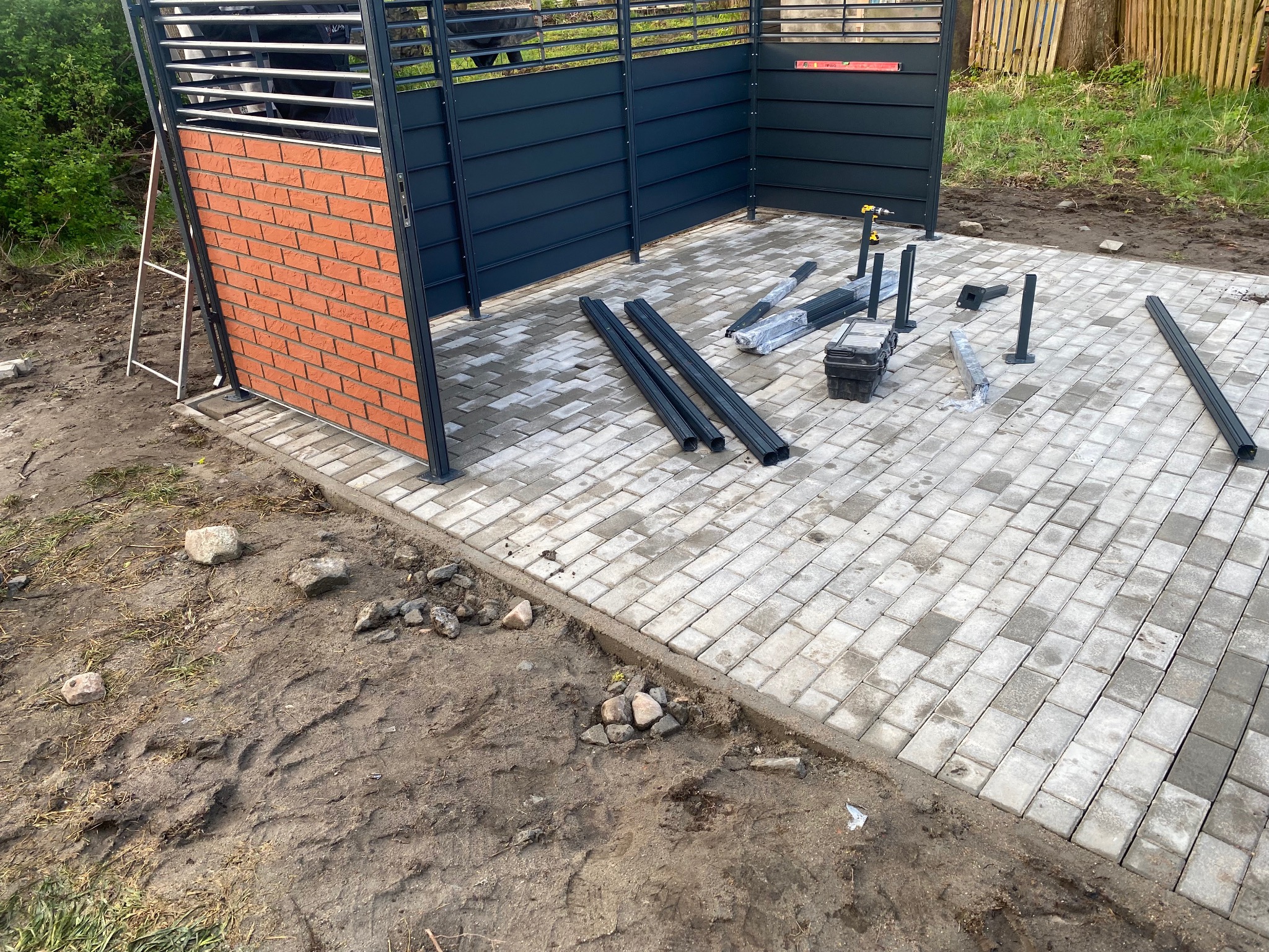 Montaż szarej, metalowej konstrukcji z elementami ceglanymi na jasnej kostce brukowej; widoczne narzędzia i elementy montażowe.