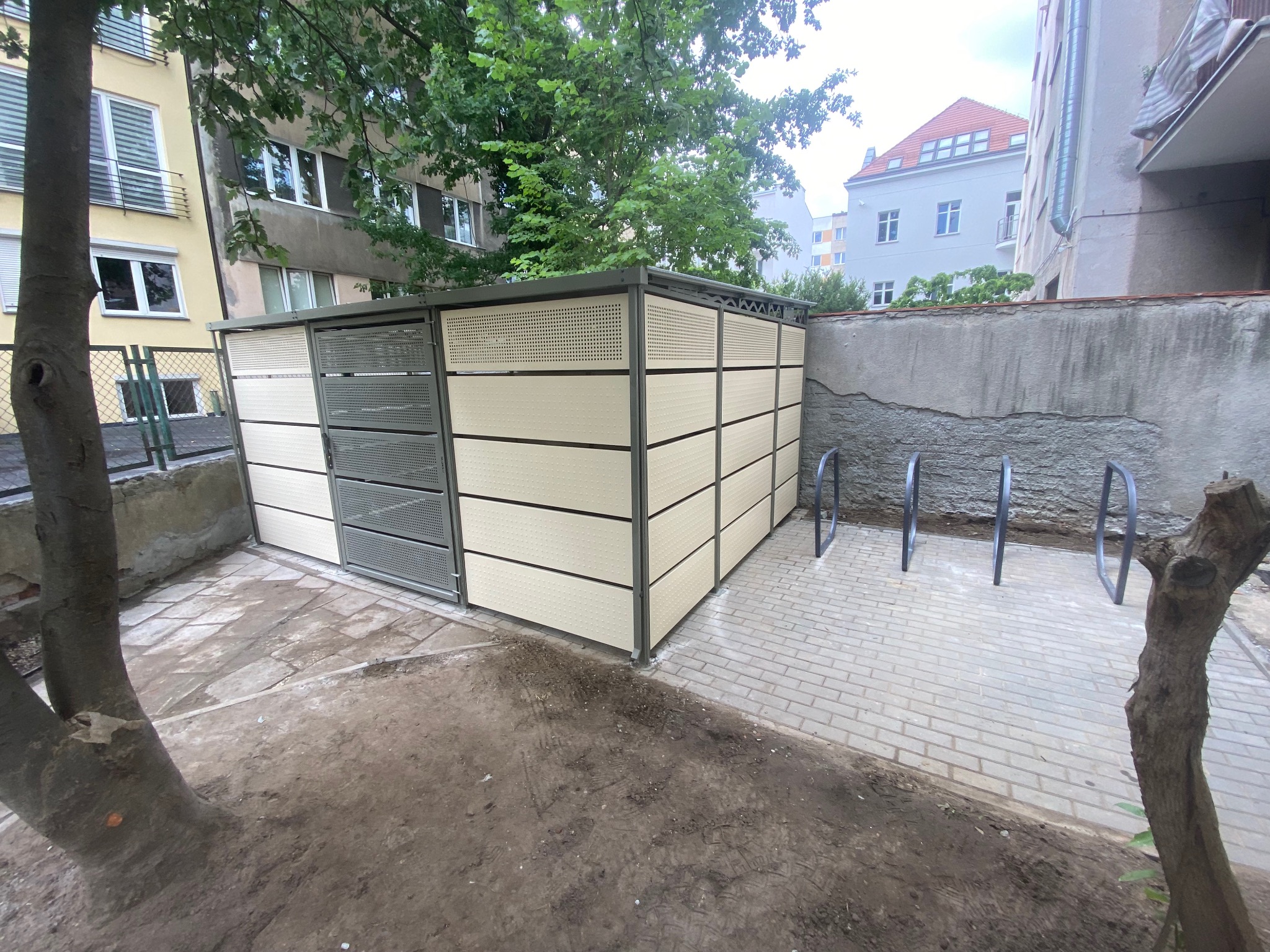 Zadaszona wiata rowerowa z perforowanymi ścianami w odcieniach szarości i beżu, ustawiona na brukowanym terenie obok drzewa i ściany budynku.