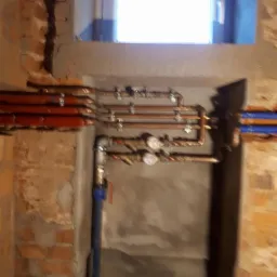 Instalacja grzewcza z miedzianymi rurami i manometrami w pomieszczeniu technicznym w Wieliczce, widoczne fragmenty ceglanego muru i okno.