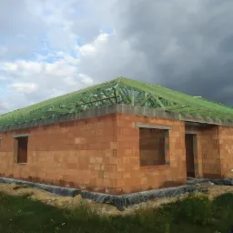 Surowy stan budynku jednorodzinnego z cegły w Pabianicach, z widoczną konstrukcją dachu pokrytą zieloną folią, na tle pochmurnego nieba.