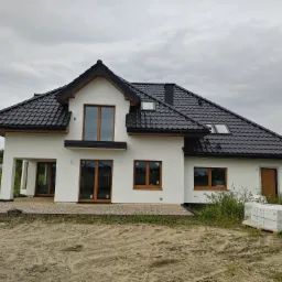 Nowoczesny dom z białymi ścianami i ciemnym dachem z dachówki ceramicznej, widoczne okna z drewnianymi ramami, palety z materiałami budowlanymi przed budynkiem, trawa i piasek na pierwszym planie.