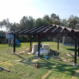 Montaż drewnianej konstrukcji altany w ogrodzie, widoczne narzędzia i materiały budowlane, w tle plac zabaw i drzewa.