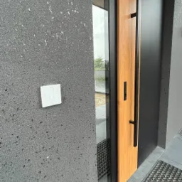 Nowoczesne wejście do domu z drewnianymi drzwiami z czarnymi okuciami, szarą elewacją z zamontowanym panelem dotykowym alarmu oraz wycieraczką.