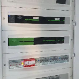 Otwarta biała skrzynka rozdzielcza z modułami Loxone, zabezpieczeniami Hager i wyłącznikiem głównym, widoczne diody sygnalizacyjne.