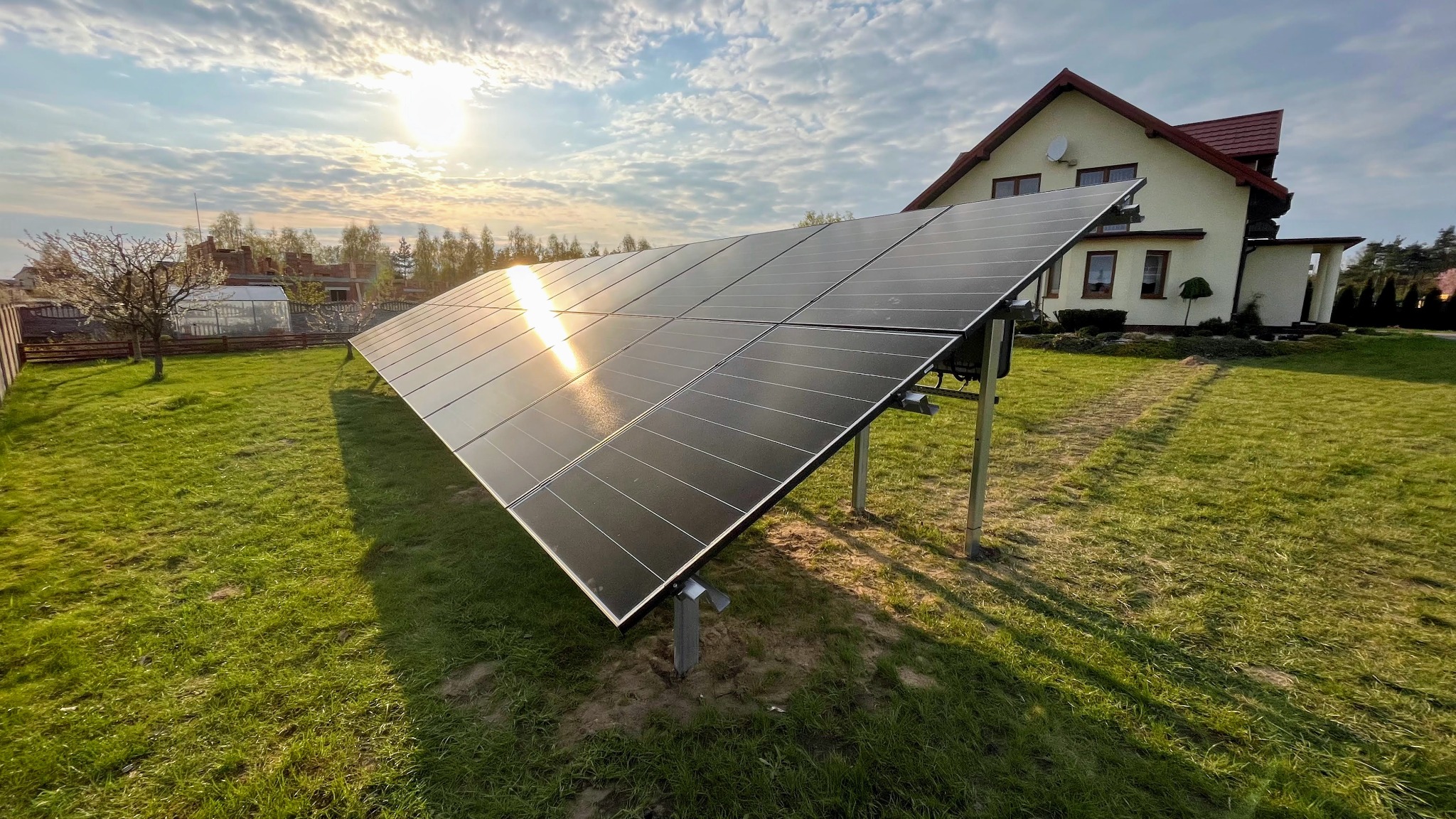 Panele fotowoltaiczne na trawniku przed domem jednorodzinnym, odbijające promienie zachodzącego słońca, widoczne metalowe podpory.