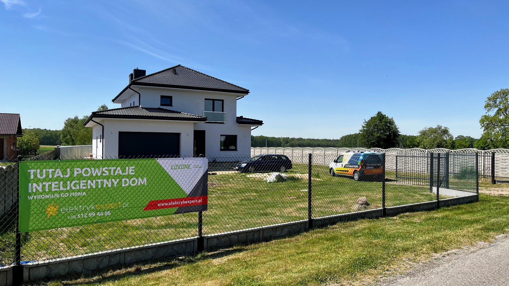 W Pajęcznie powstaje nowoczesny dom z systemem inteligentnego zarządzania, o czym informuje baner reklamowy firmy elektrykexpert.pl, na posesji zaparkowany firmowy samochód z logotypem fotowoltaiki.