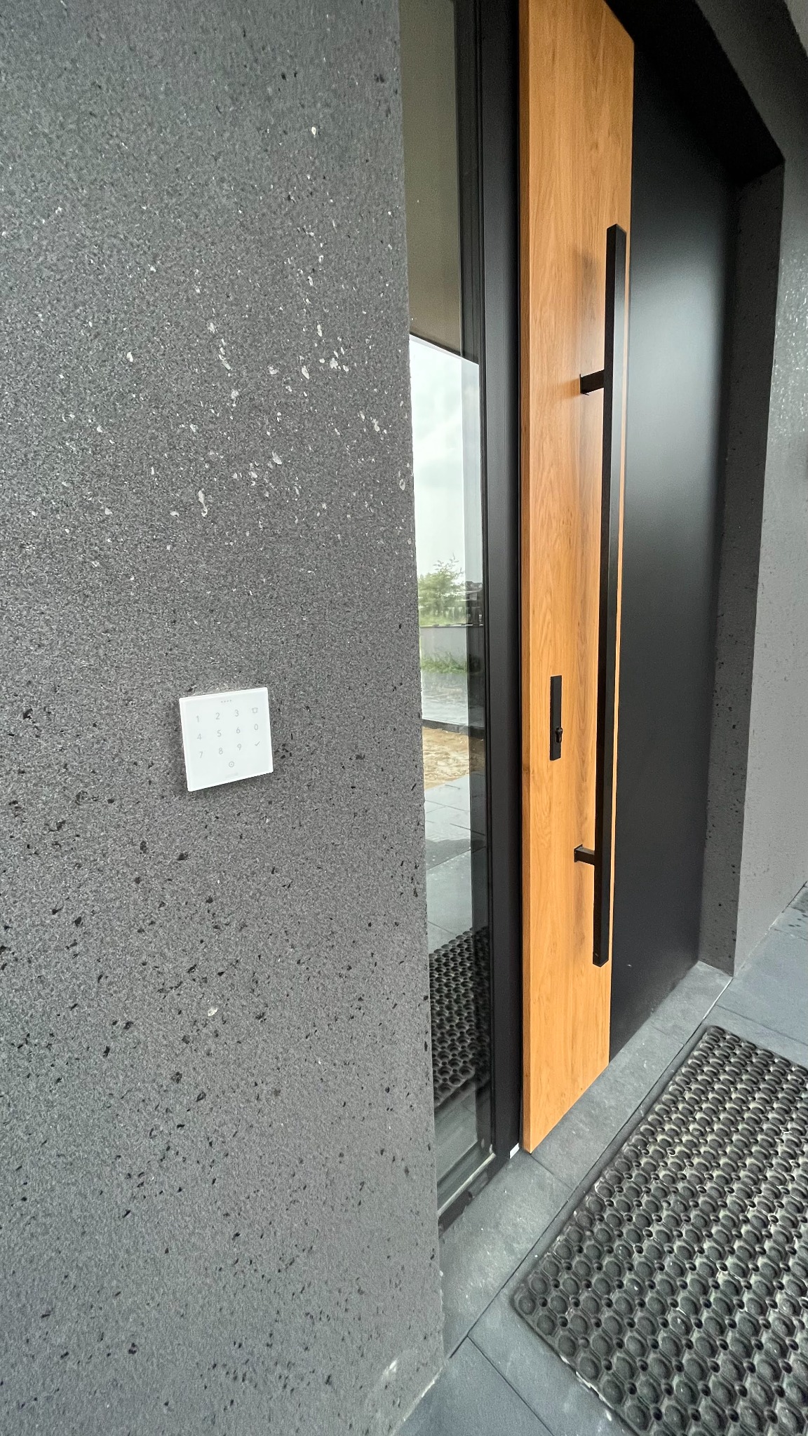 Nowoczesne wejście do domu z drewnianymi drzwiami z czarnymi okuciami, szarą elewacją z zamontowanym panelem dotykowym alarmu oraz wycieraczką.