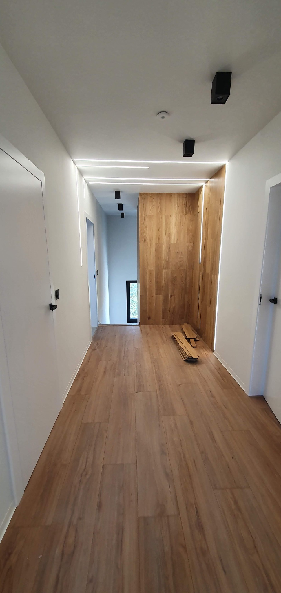 Nowoczesny korytarz z podświetlanymi listwami LED w suficie i ścianach, drewnianą okładziną ściany i brązowymi panelami podłogowymi. Na podłodze leżą docięte panele.