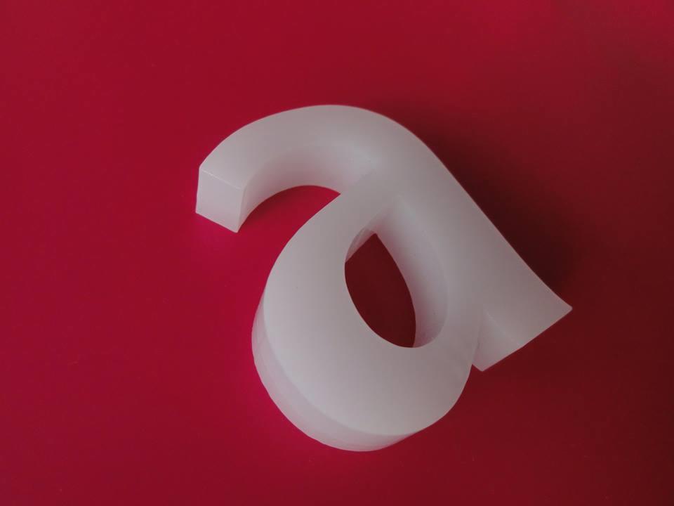 Biała, przestrzenna litera 'a' wykonana z tworzywa sztucznego, ustawiona pod kątem na gładkim, czerwonym tle.