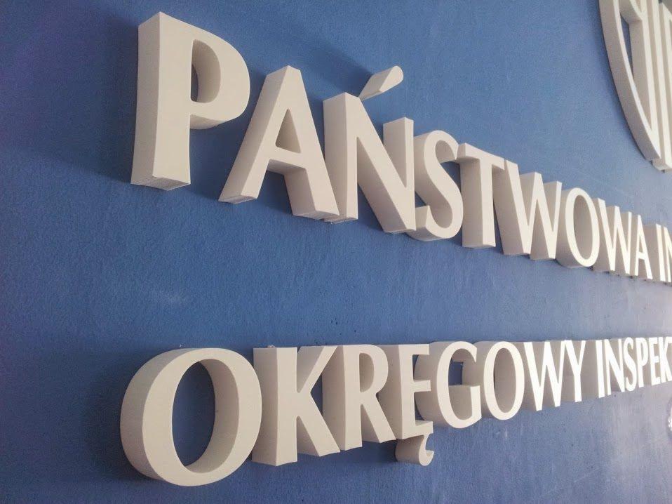 Przestrzenne, białe litery tworzące napis 'PAŃSTWOWA INSPEKCJA OKRĘGOWY INSPEKTOR' na niebieskiej ścianie, widok z boku.