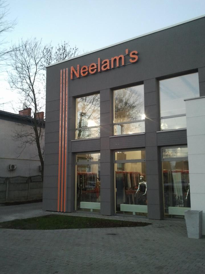 Elewacja budynku z szyldem 'Neelam's' z pomarańczowymi paskami, widoczne okna wystawowe z manekinami w środku, szara kostka brukowa na chodniku.
