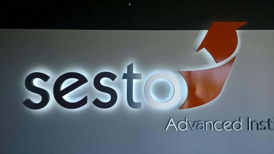 Podświetlane logo firmy 'sesto' z pomarańczowym akcentem w kształcie strzałki, zamontowane na ścianie w biurze.