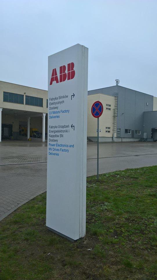 Pylon informacyjny z logo ABB i nazwami fabryk oraz kierunkami, ustawiony na tle budynków fabrycznych i znaku zakazu parkowania.