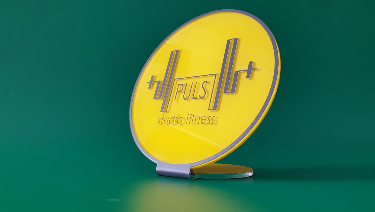 Żółty, okrągły szyld z szarym obramowaniem i napisem 'PULS studio fitness' na zielonym tle, podświetlony z góry.