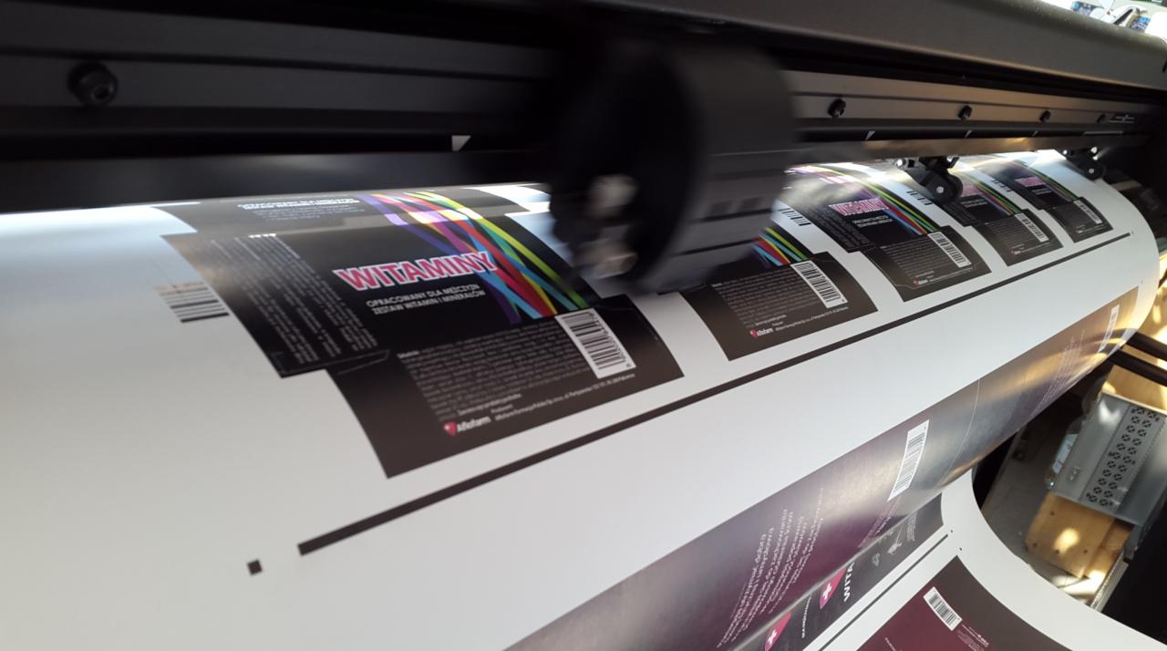 Drukarka wielkoformatowa w trakcie drukowania etykiet na rolce papieru, widoczne napisy 'Witaminy' i kolorowe paski.