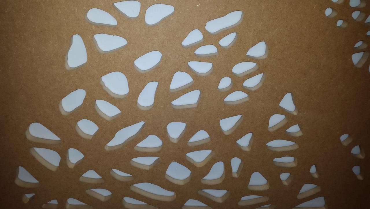 Brązowa płyta z wyciętymi nieregularnymi otworami, przez które prześwituje białe tło.