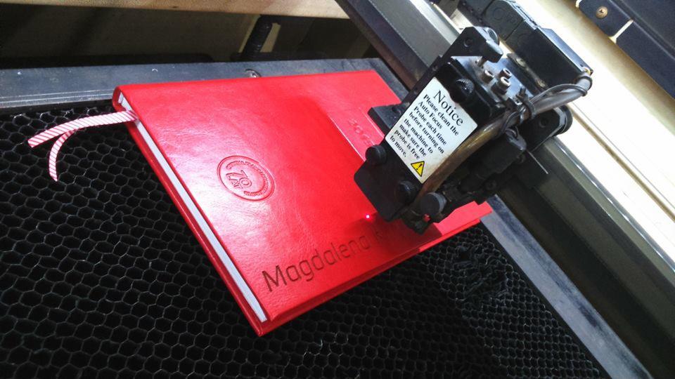 Czerwony notatnik z imieniem 'Magdalena' grawerowanym laserowo, umieszczony na platformie maszyny do grawerowania, z widocznym laserem w trakcie pracy.