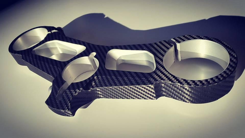 Szczegółowy widok mostka kierownicy motocykla wykonanego z włókna węglowego z aluminiowymi elementami mocującymi, oświetlony z boku, tworzący cień.