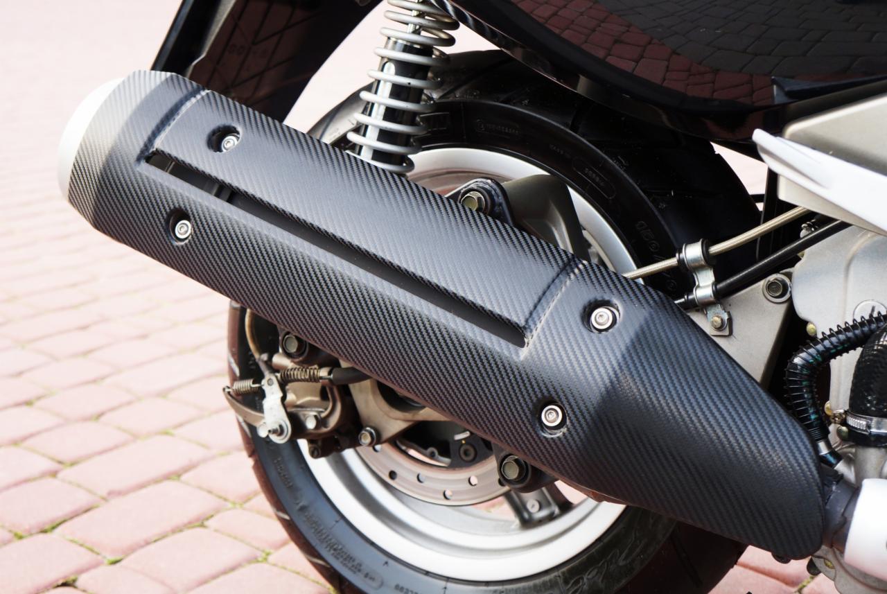 Czarny, teksturowany osłona wydechu skutera z widocznymi śrubami mocującymi, na tle fragmentu koła, amortyzatora i kostki brukowej.