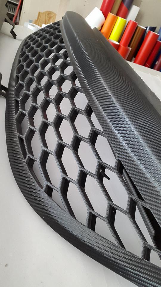 Element karoserii samochodowej, prawdopodobnie grill, pokryty folią imitującą włókno węglowe. Widoczne rolki kolorowej folii w tle.