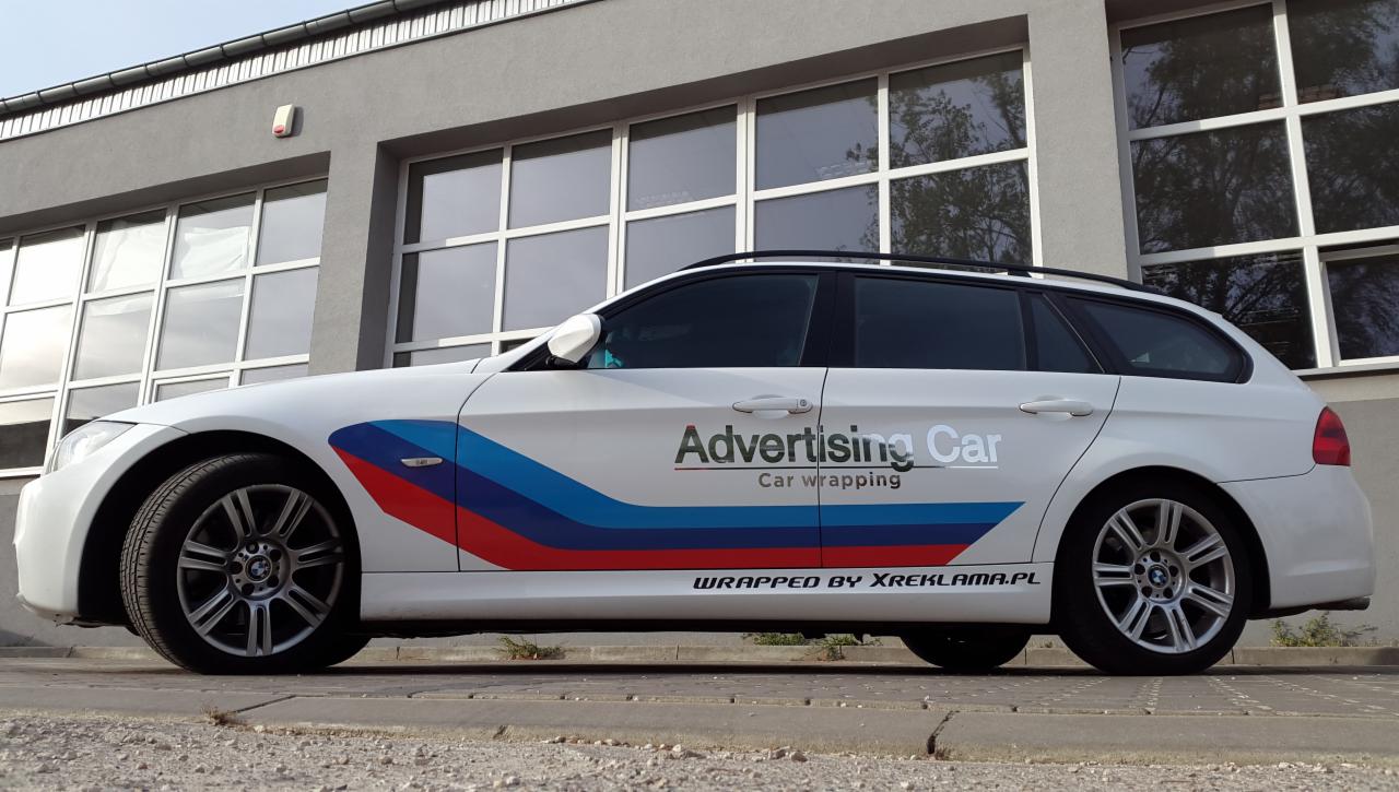 Białe BMW kombi z reklamą na karoserii, z napisem 'Advertising Car' i trójkolorowymi paskami, zaparkowane przed szarym budynkiem z dużymi oknami.