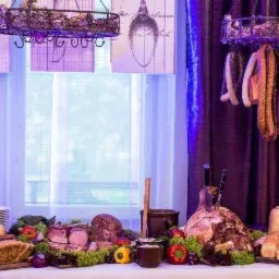 Bogato zastawiony stół wiejski z wędlinami, pieczywem, warzywami i dekoracjami, w tle okno z zasłonami i wiszące wędliny