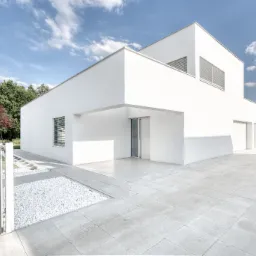 Nowoczesny, minimalistyczny dom w Tychach z białymi ścianami, jasną kostką brukową i dekoracyjnym żwirem, widoczny fragment ogrodu z drzewami i pole kukurydzy w tle, brama wjazdowa z metalowymi...